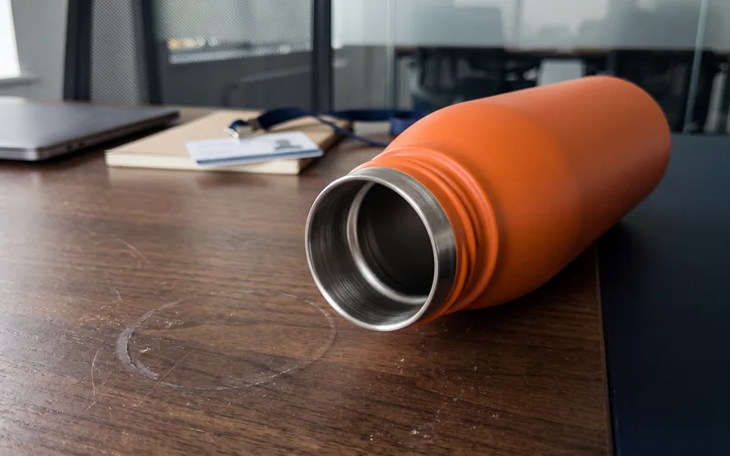 Garrafa térmica laranja fosca aberta e vazia sobre a mesa de nogueira de uma sala de reunião, deitada de lado com a boca voltada para a câmera: vê-se a espessura da parede dupla — a casca laranja por fora e o anel de inox escovado por dentro. Ao fundo, desfocados, um notebook fechado, um caderno e um crachá.
